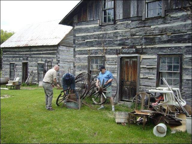 Scott Adkins, Brian Adkins and auctioneer Kenny Lindsay (from left to right) assemble an antique wagon wheel at the  Rustic Village Museum in Waltz. The museum and a general store (building at far left), both made of logs was sold at an auction handled by Ken Lindsay of American Eagle Auction & Appraisal Company.